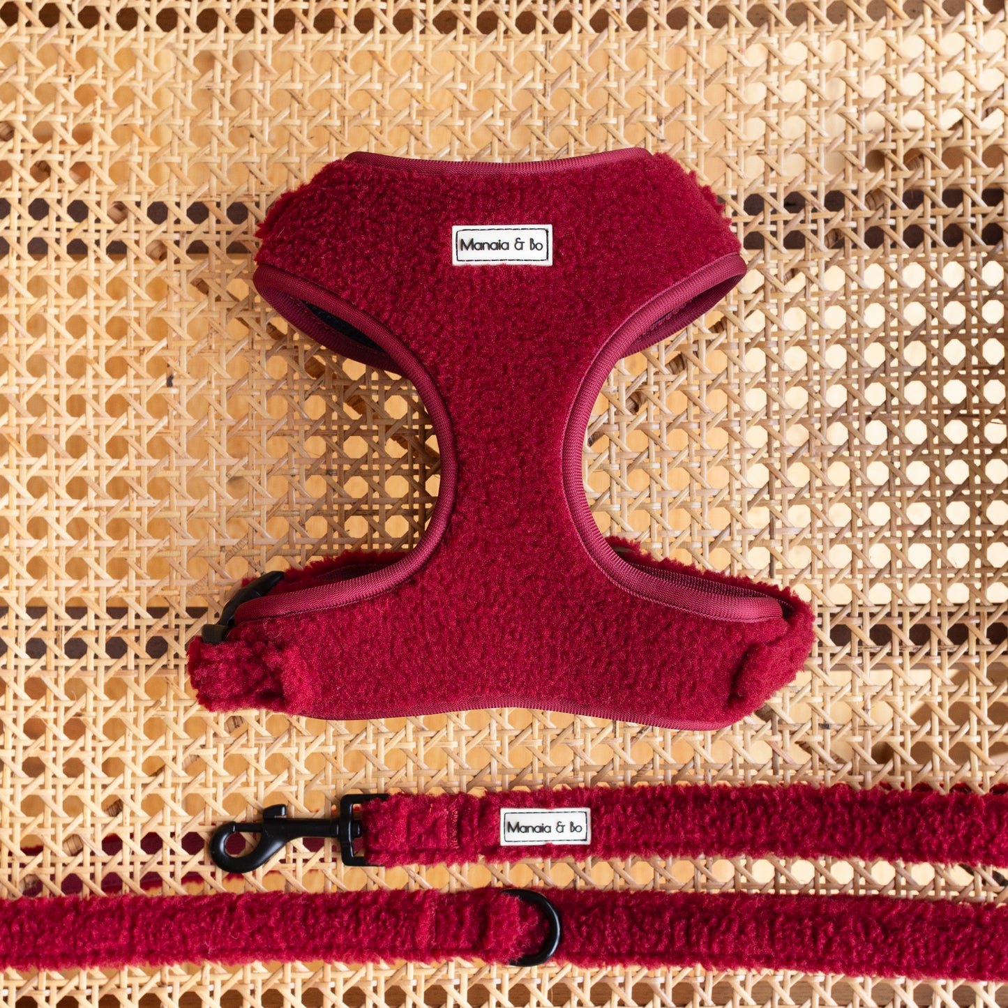 The Bordeaux Teddy Harness, Lead & Collar set in rich red teddy fabric with black hardware, displayed on a woven wicker surface.