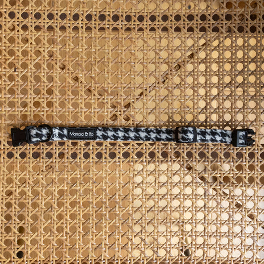 The Classic Houndstooth Dog Collar, featuring black plastic buckles and Monica & Jo text, rests on a woven rattan surface—offering a stylish, adjustable accessory for your pet.
