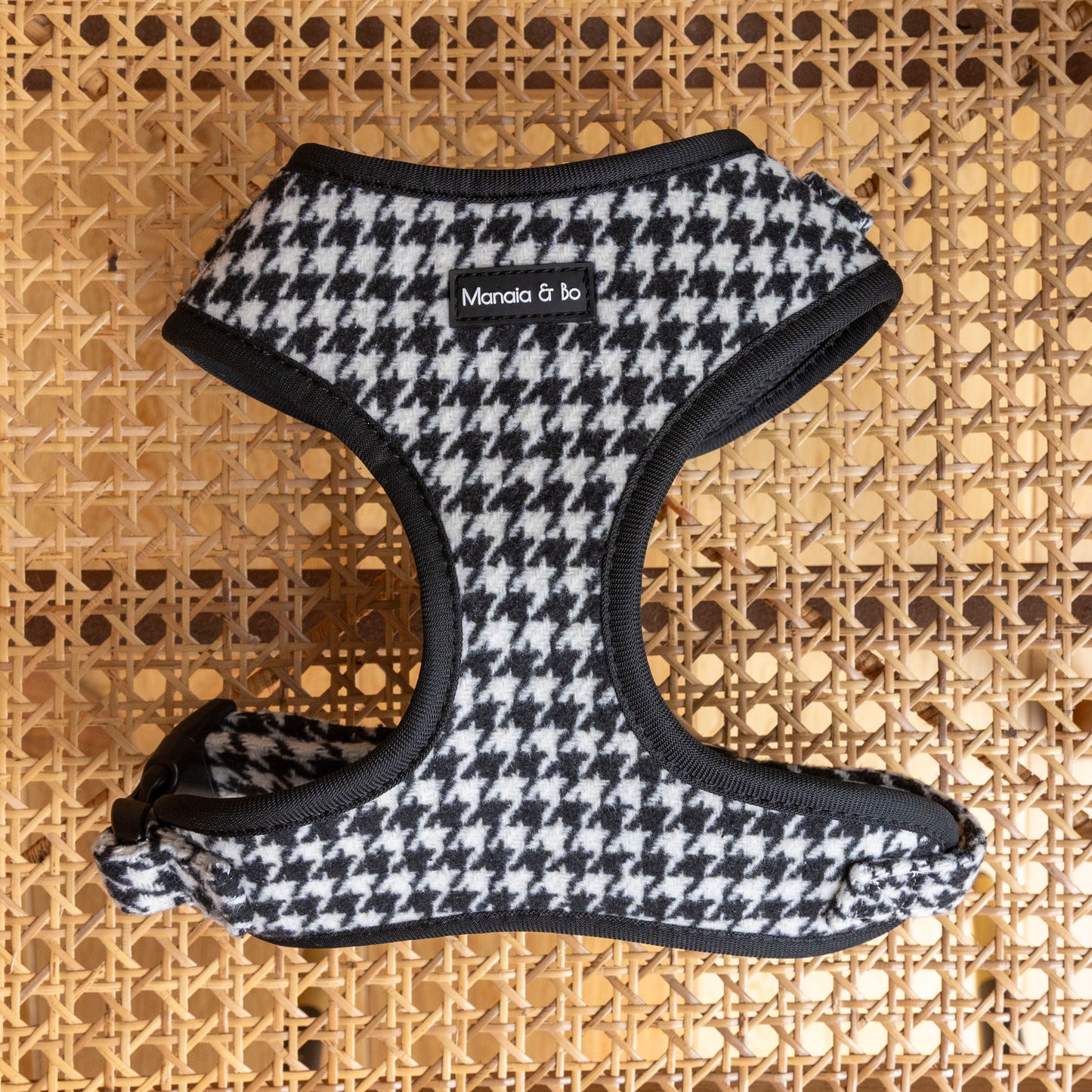 The Classic Houndstooth Harness, Lead & Collar in black and white houndstooth tweed with black trim, by Manola & Bo, is shown on a woven cane surface—ideal to pair with the matching lead or adjustable collar.