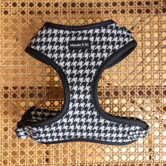 The Classic Houndstooth Harness, Lead & Collar in black and white houndstooth tweed with black trim, by Manola & Bo, is shown on a woven cane surface—ideal to pair with the matching lead or adjustable collar.