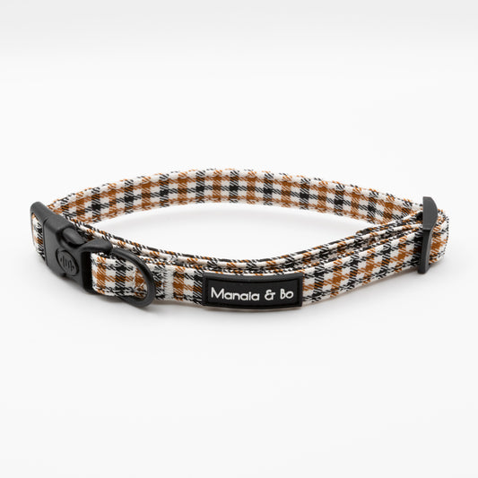 The Cocoa Heritage Lite Dog Collar features a plaid pattern in black, white, and brown, an adjustable fit with lockable buckle, metal ring, and a Manala & bo label on the side.