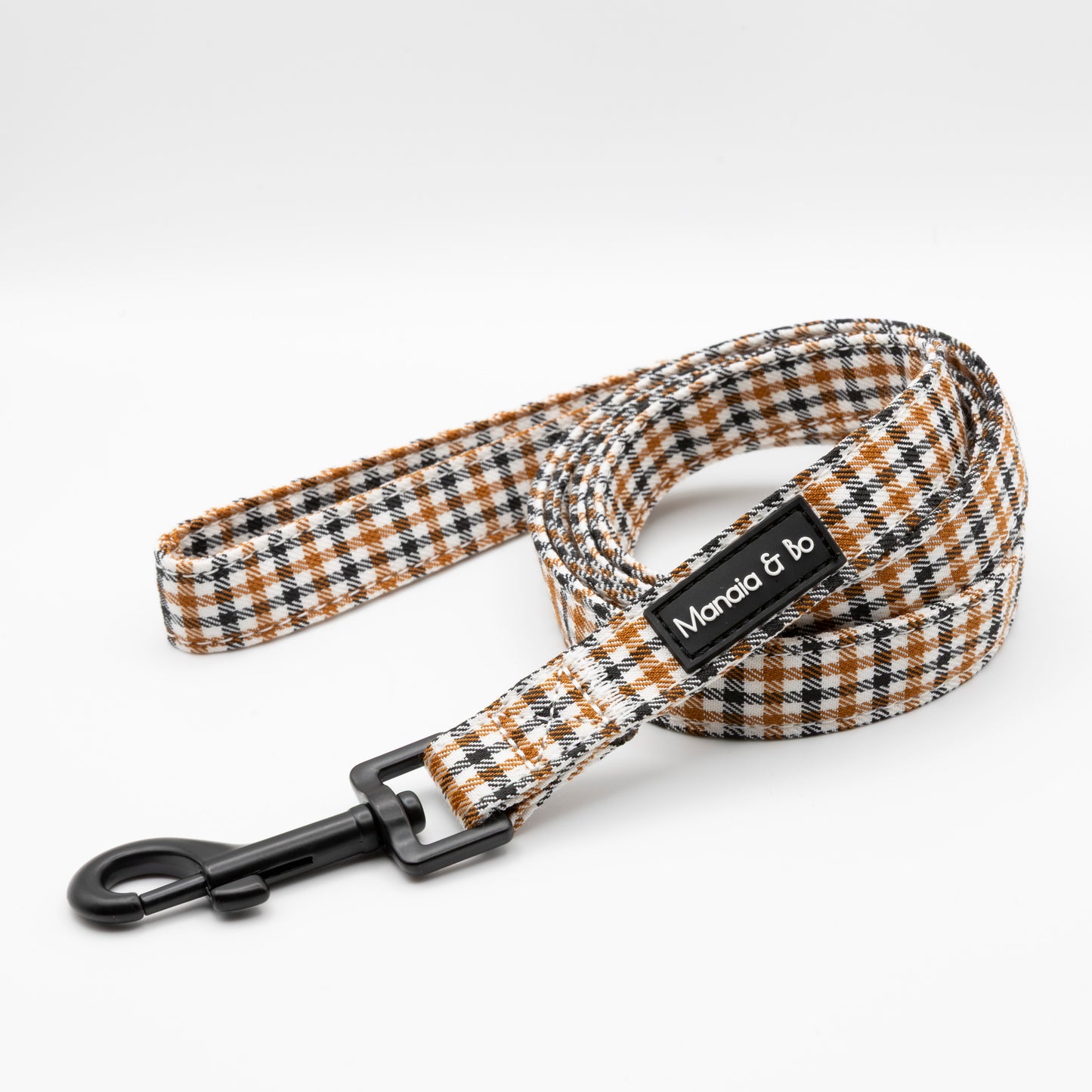 The Cocoa Heritage Lite Dog Lead is a coiled leash with a black, white, and brown plaid pattern, a black metal clasp, and a Monica & Bo label on a white background, inspired by our heritage check dog harness.