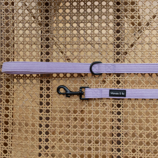 The Lavender Cord Lead featuring a a black fastening hook and d-ring, rests on a woven wicker surface. The lead is perfect for pairing with our comfortable pet harness as the ideal walking set.
