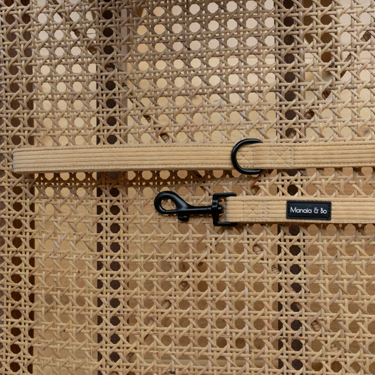 The Mellow Cord Dog Lead, featuring a beige design with a black metal hook and D-ring, showcases quality materials. the lead lies flat on a cane chair with an elegant woven pattern and circular gaps.