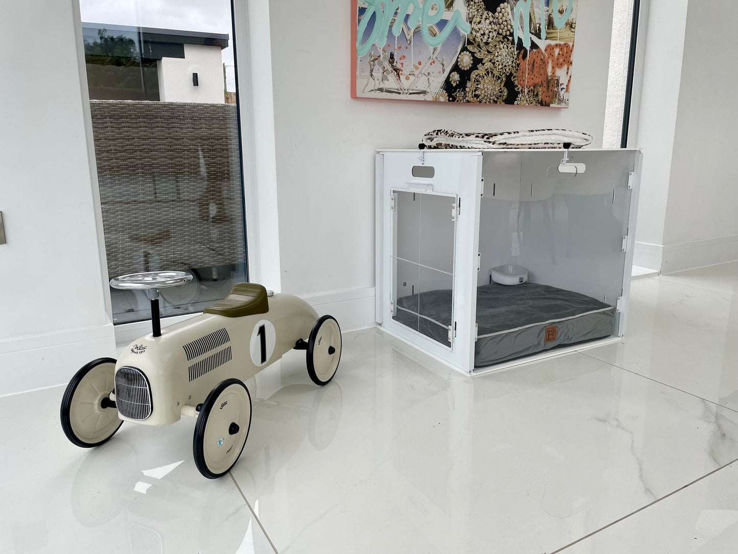 A cream-colored toy race car with the number 1 sits on a shiny white floor beside a minimalist Petpen Crate featuring a gray cushion, in a bright room with large windows.