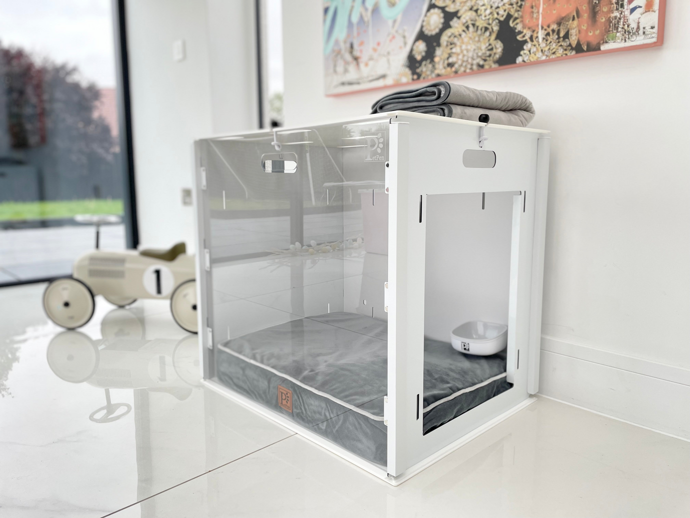 The Petpen Crate, featuring a glass door, sits in a bright room with a gray cushion and water bowl inside for crate training. A folded blanket rests on top, while a toy car lies nearby on the shiny floor.