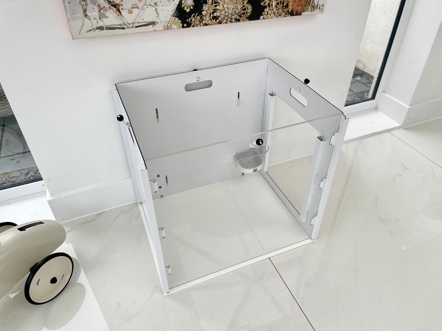The Petpen Crate, a transparent minimalist pet crate with a small door and ventilation holes, is placed on a white tiled floor by a window. A white toy car is partly visible on the left, enhancing the clean, modern look.