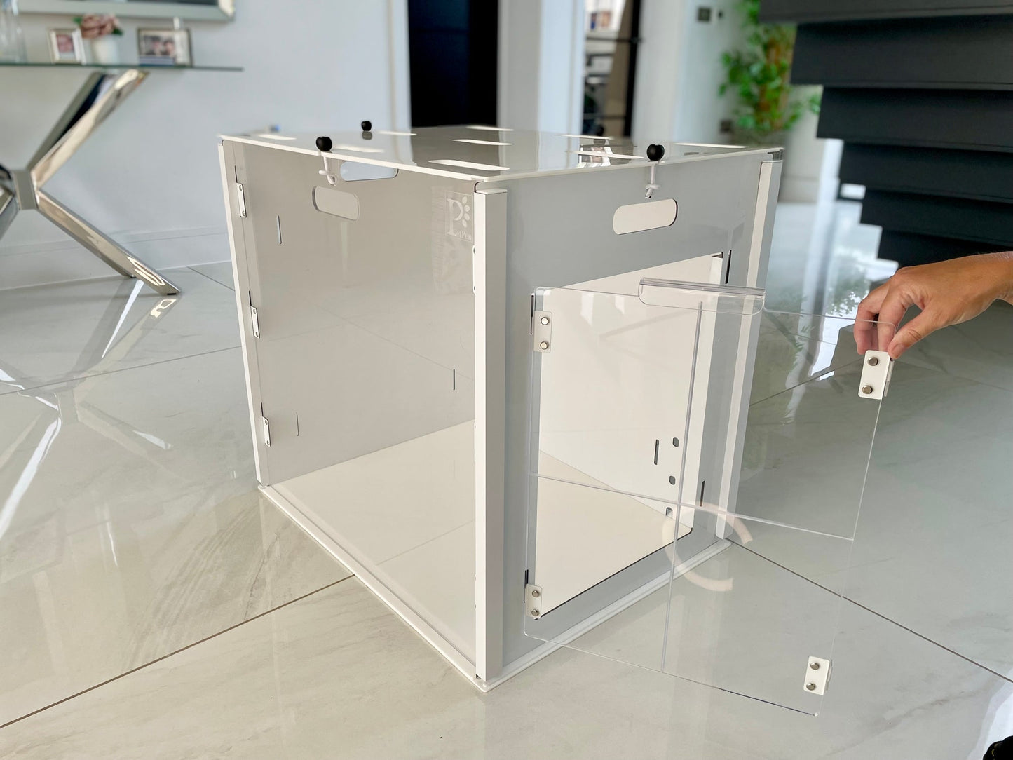 A hand opens the hinged door of a transparent Petpen Crate, which sits on a shiny tiled floor in a modern, bright room with contemporary furniture in the background.