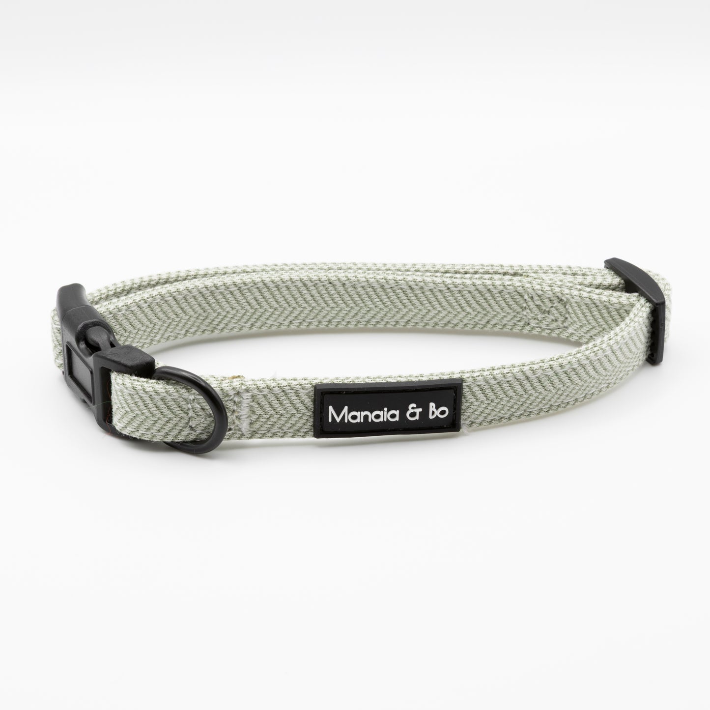 The Pine Tweed Dog Collar is light gray pine tweed with a black plastic buckle, metal D-ring, and a black and white Manala & bo label. Displayed in a circular shape on a white background, this collar is adjustable.