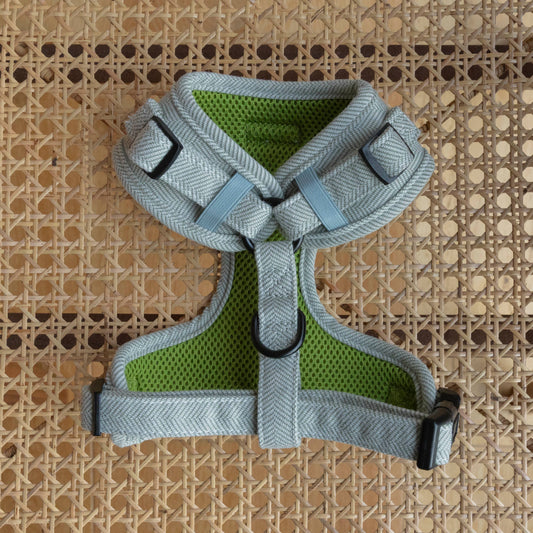 The Pine Tweed Harness sits on a woven rattan surface, showcasing its gray and green herringbone pattern. This adjustable dog harness includes straps for a perfect fit, a back D-ring, and a padded interior for comfort.