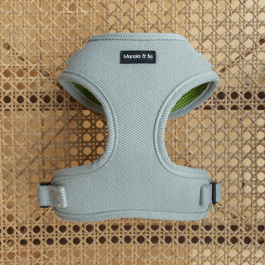 The Pine Tweed Dog Harness in sage green features a herringbone pattern and sits on a wicker background. This adjustable harness offers quality pet materials, an ergonomic design with two leg openings, and adjustable straps for comfort.