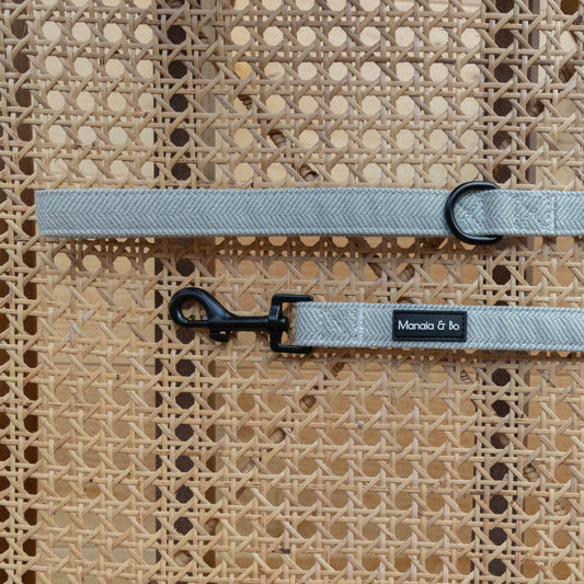 A light grey Pine Tweed Dog Lead with a black metal clip rests on a woven cane surface. Made from quality materials, it has a black ring near one end.