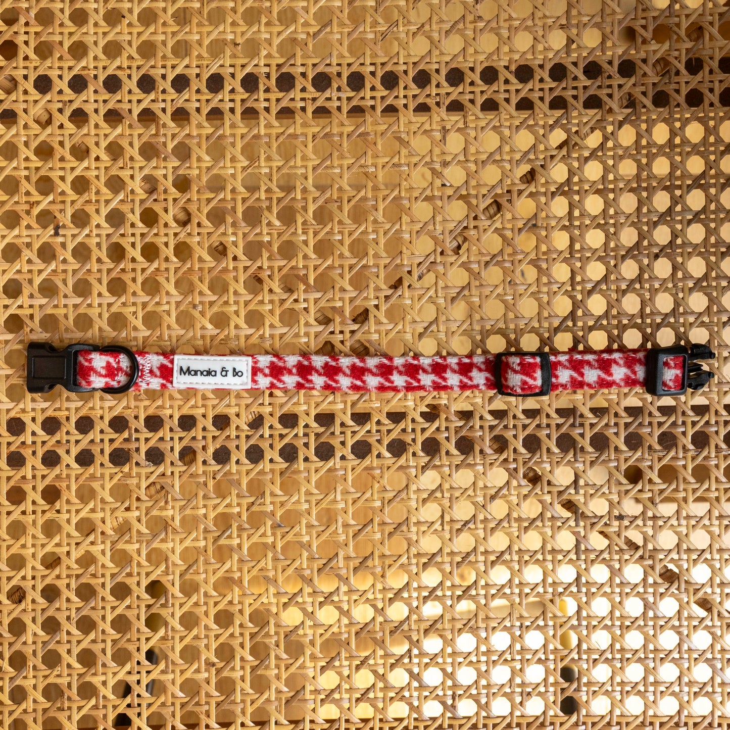 The Scarlet Houndstooth Dog Collar, featuring a red and white houndstooth pattern with a black buckle and adjustable clips, is ideal for small to medium dogs and is shown lying flat on a woven cane chair seat.