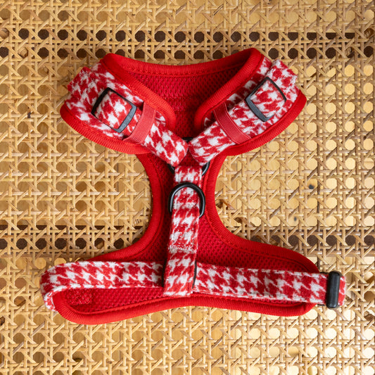 The Scarlet Houndstooth Dog Harness, featuring adjustable straps and black buckles, is displayed on a woven cane chair seat.