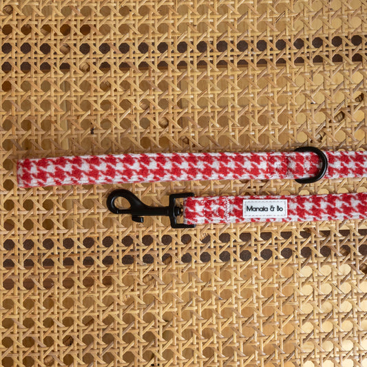 The Scarlet Houndstooth Dog Lead, featuring a red and white tweed pattern with a black metal clip and ring, rests on woven wicker. A white Mandai & to label adds a stylish touch to this fixed-length accessory.
