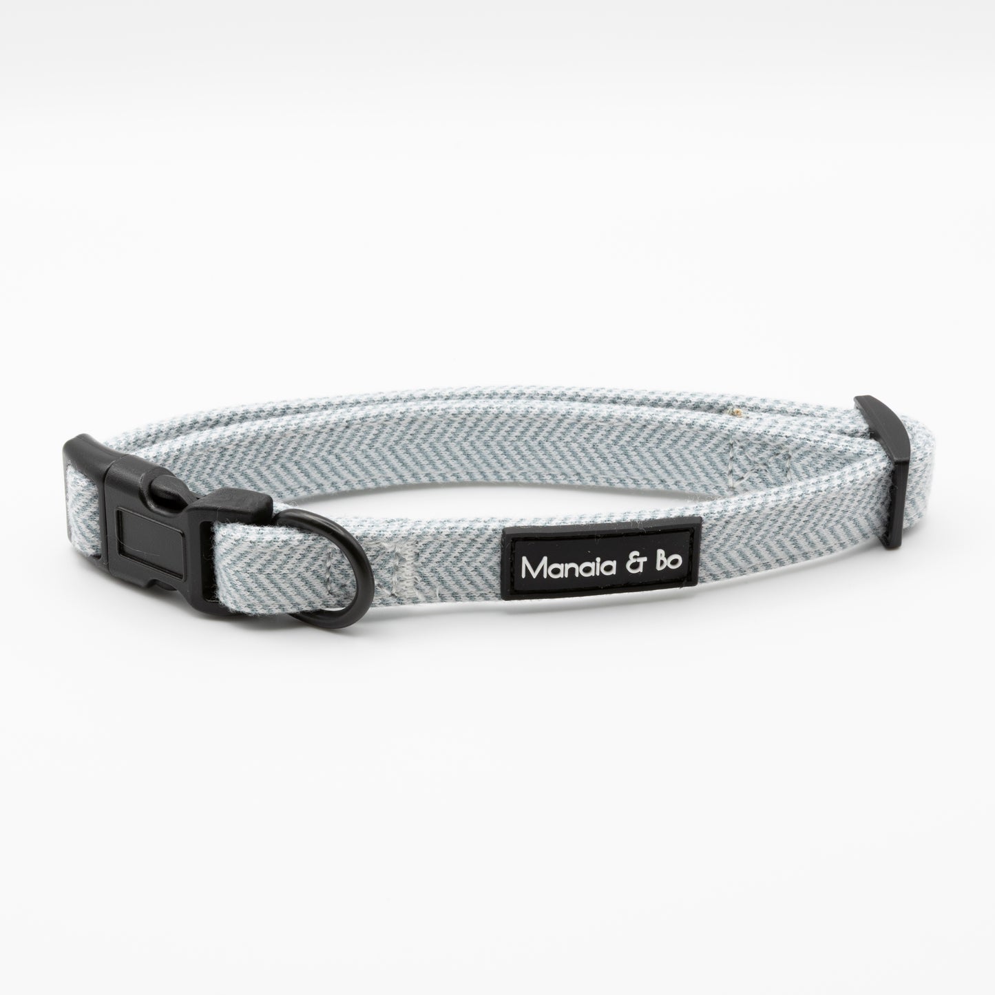 The Sky Tweed Dog Collar features light gray herringbone fabric, a black plastic buckle, and metal D-ring. Adjustable for small to medium dogs, it has a black "Manala & Bo" label on white.
