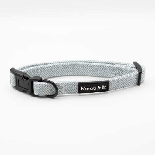 The Sky Tweed Dog Collar features light gray herringbone fabric, a black plastic buckle, and metal D-ring. Adjustable for small to medium dogs, it has a black "Manala & Bo" label on white.