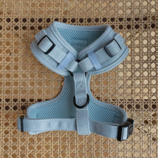 A light gray Sky Tweed Harness lies on a rattan surface, the harness is face down, highlighting the light blue breathable mesh. It has adjustable straps, black buckles, a D-ring for leash attachment, and features a soft, breathable mesh interior with a herringbone-patterned exterior.
