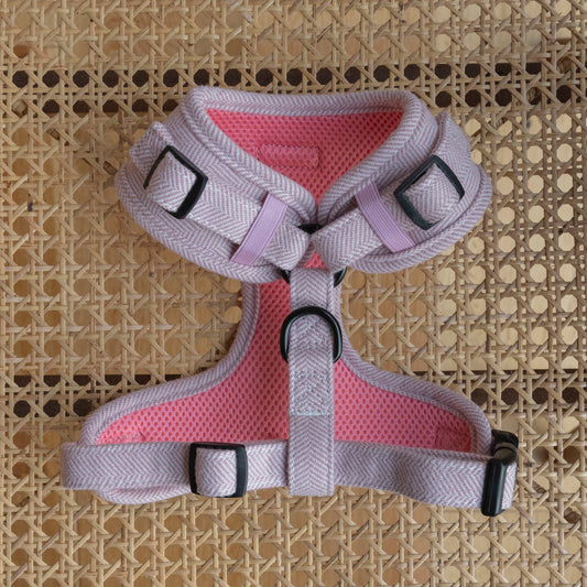The Tea Rose Tweed Harness, pictured from the reverse side, in pink and purple with adjustable black buckles, lies on a woven rattan surface. Crafted from quality materials, it includes a handle and metal rings for leash attachment. The pink breathable mesh is highlighted in the image