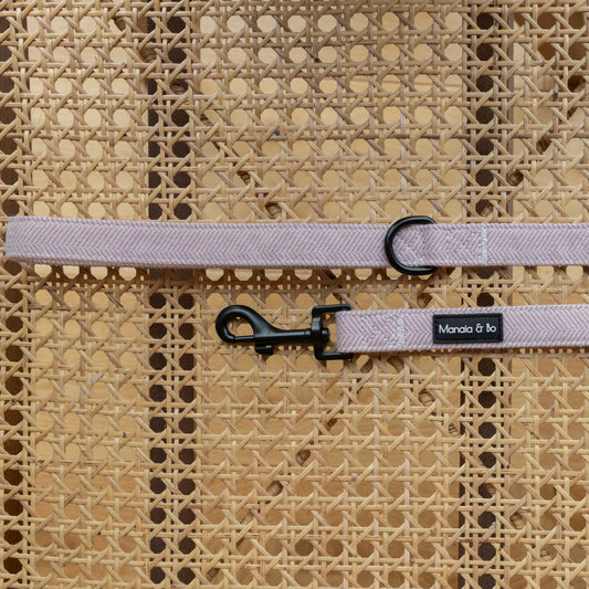 The Tea Rose Tweed Dog Lead, a pink woven leash with a black clasp and D-ring, rests elegantly on a wicker chair. Crafted from quality materials, this fixed-length lead is a perfect addition to a matching adjustable harness or collar