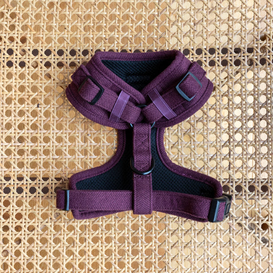 The Wild Plum Tweed Dog Harness, featuring black buckles and adjustable straps, is displayed on a woven rattan surface—a stylish and comfortable purple harness for dogs.