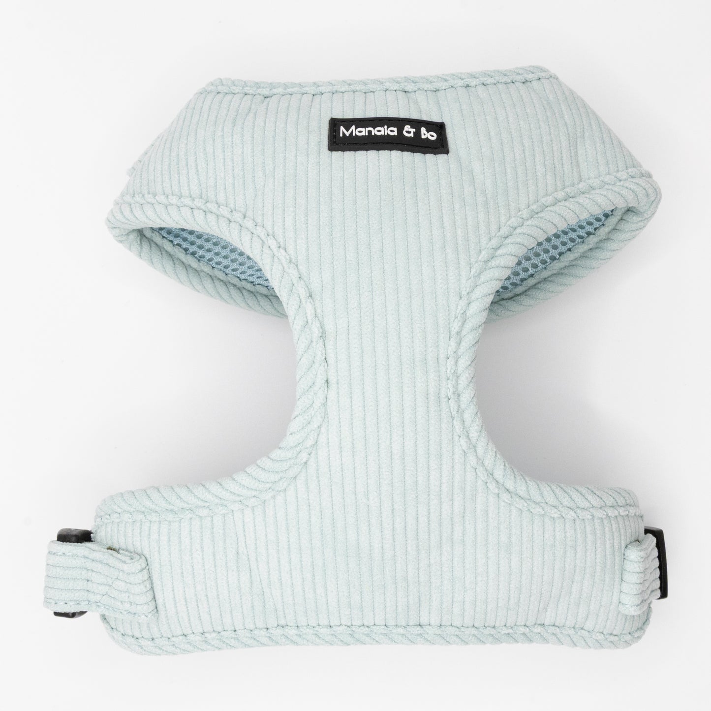 The Aqua Cord Dog Harness is light blue with ribbed aqua fabric and a black and white Manala & Bo label on the front. It has a buckle, adjustable strap for comfort, and is shown against a white background.