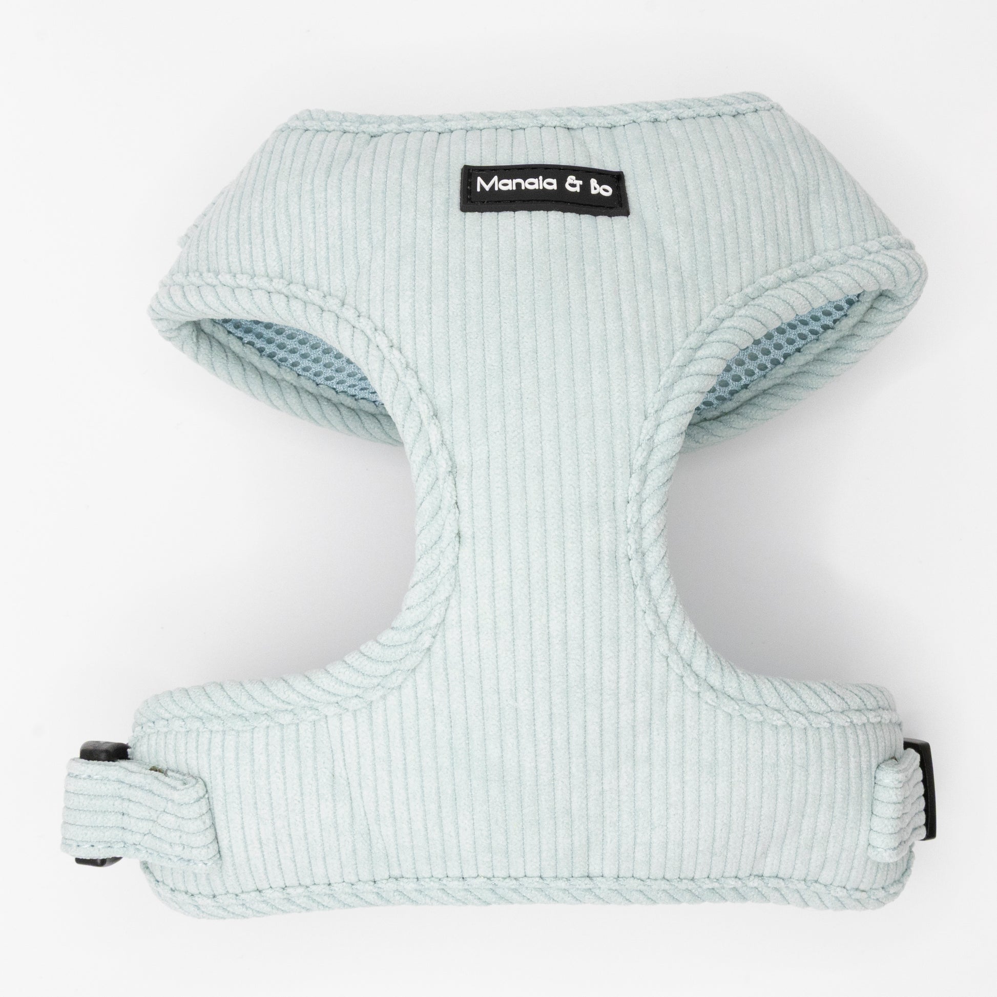 The Aqua Cord Dog Harness is light blue with ribbed aqua fabric and a black and white Manala & Bo label on the front. It has a buckle, adjustable strap for comfort, and is shown against a white background.