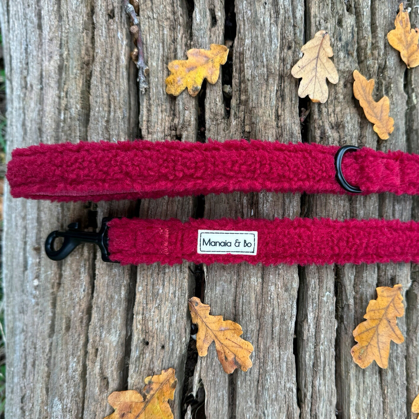 Bordeaux (burgundy) teddy material dog lead laying on a tree trunk from top showing both ends. Comfortable material with black metal d-ring by the lead handle and black metal swivel snap hook.