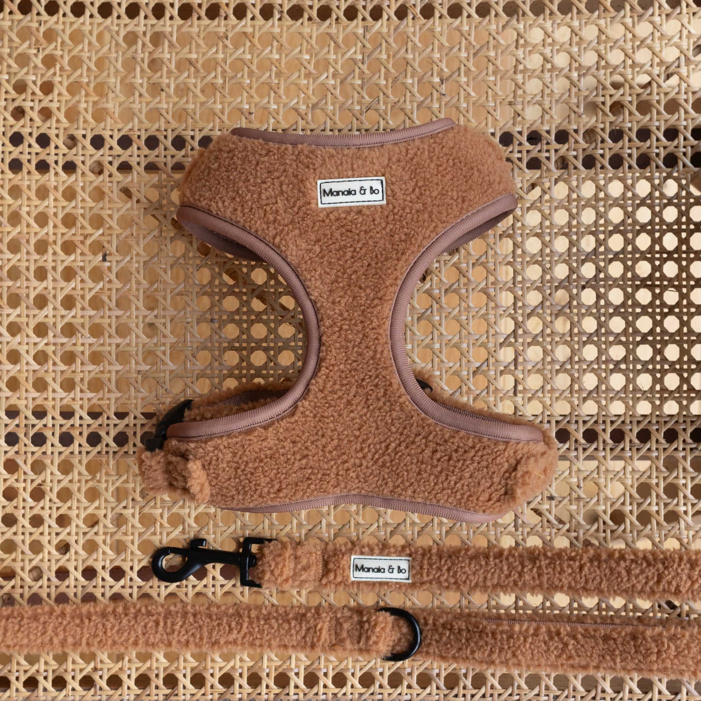 A Caramel teddy fluffy harness with the Caramel Teddy Dog Lead is displayed on a rattan surface, featuring black fastenings and a metal swivel hook.