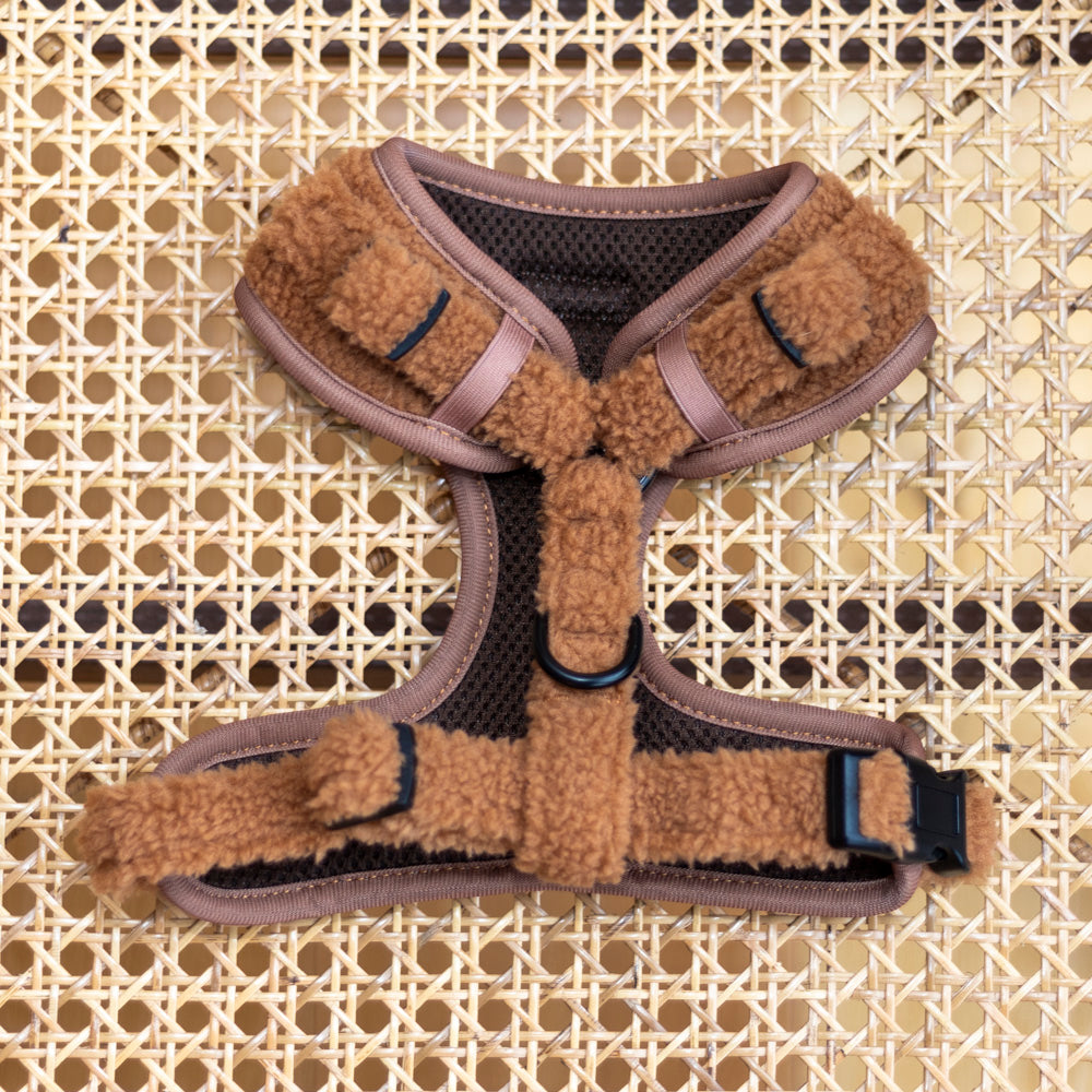 Caramel (light brown) teddy material adjustable dog harness laying on a wicker background. The harness is viewed from the back showing the dark brown mesh lining and the black plastic fastenings with the black metal fixing ring for attaching a lead.