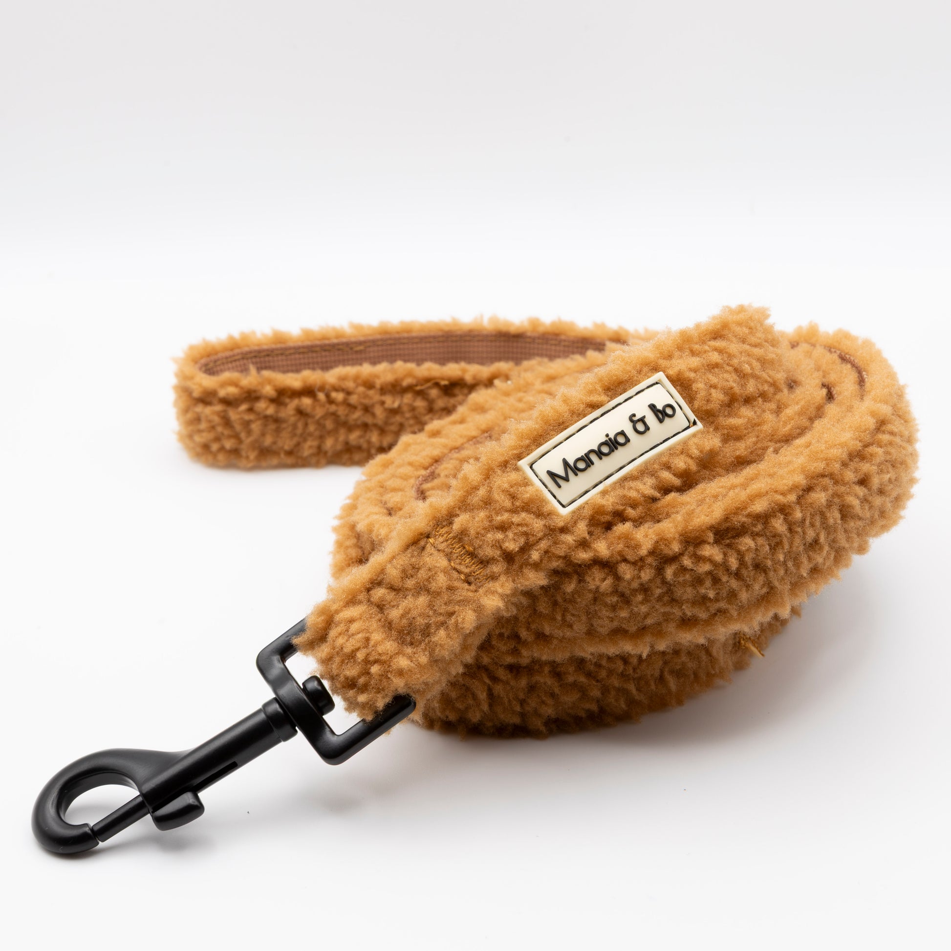 A fluffy, tan Caramel Teddy Dog Lead with a black metal clasp is coiled on a white background. Featuring a Manda & bo label, this stylish fixed length leash is perfect for your pup.