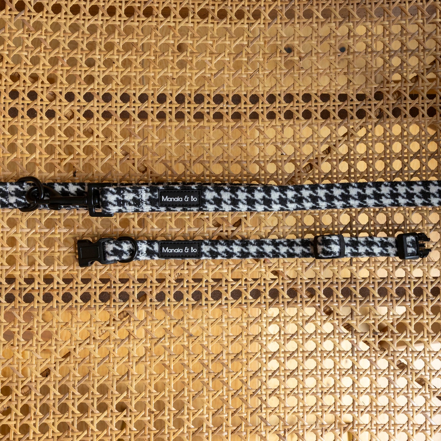 Two Classic Houndstooth Collar & Lead sets with black hardware are displayed on a woven cane surface. Each adjustable collar features a small “Monada & Co.” label.