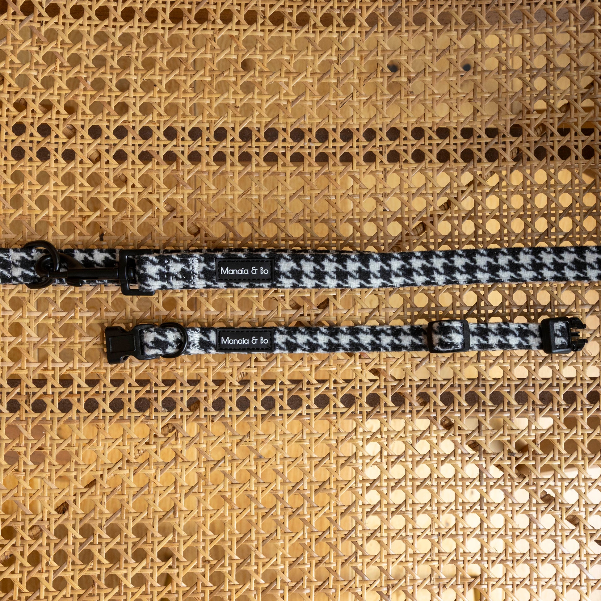 Two Classic Houndstooth Collar & Lead sets with black hardware are displayed on a woven cane surface. Each adjustable collar features a small “Monada & Co.” label.