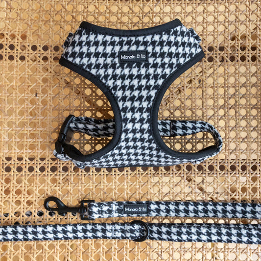 The Classic Houndstooth Harness & Lead, featuring black trim and a Manola & Io label, rests on a woven cane surface for a chic, coordinated look. This adjustable harness comes in stylish black and white houndstooth.