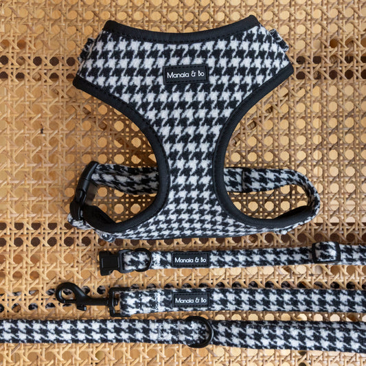 The Classic Houndstooth Harness, Lead & Collar set is displayed on a woven rattan surface, each piece featuring the Manola & Lo label.
