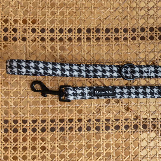 The Classic Houndstooth Harness, Lead & Collar by Manola & Bo features a black and white houndstooth-patterned dog lead with a metal clasp and ring, shown on a woven rattan surface.