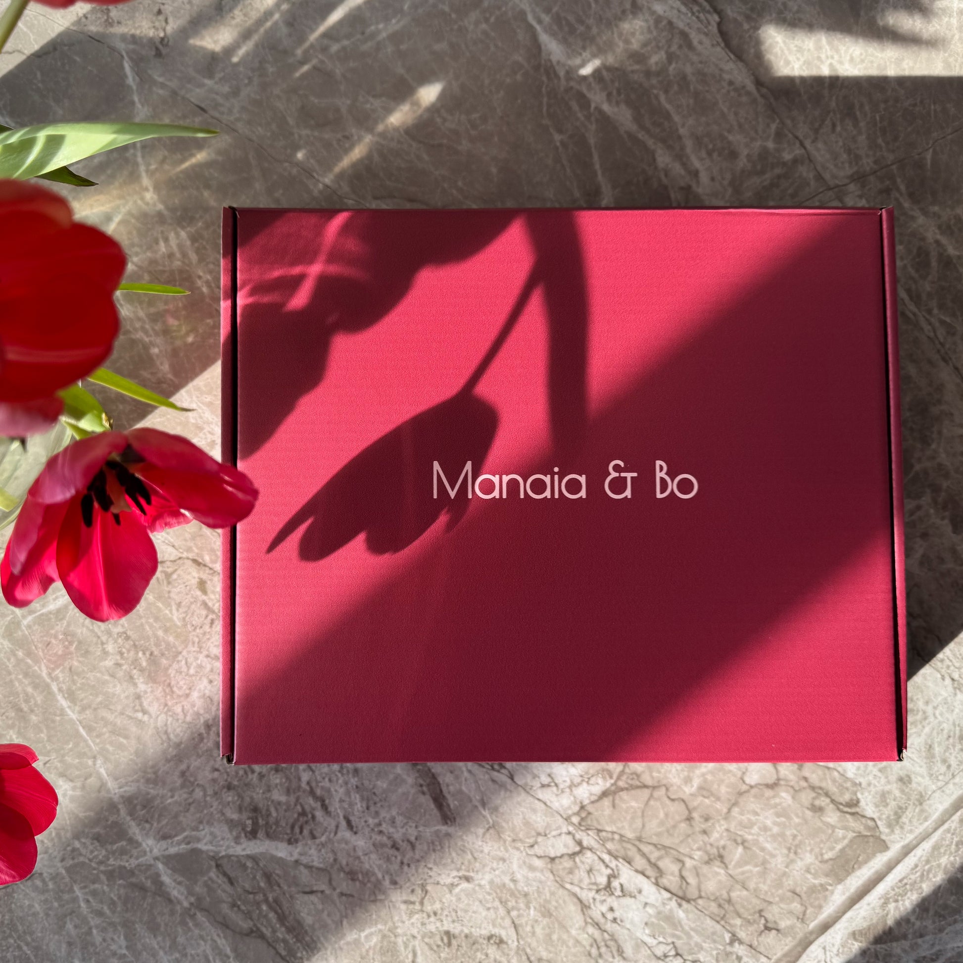 A red Manaia & Bo Gift Box sits elegantly on a stone surface, with the shadow of a tulip gracing it. Vibrant red tulips with lush green leaves enhance the charm at the left edge.