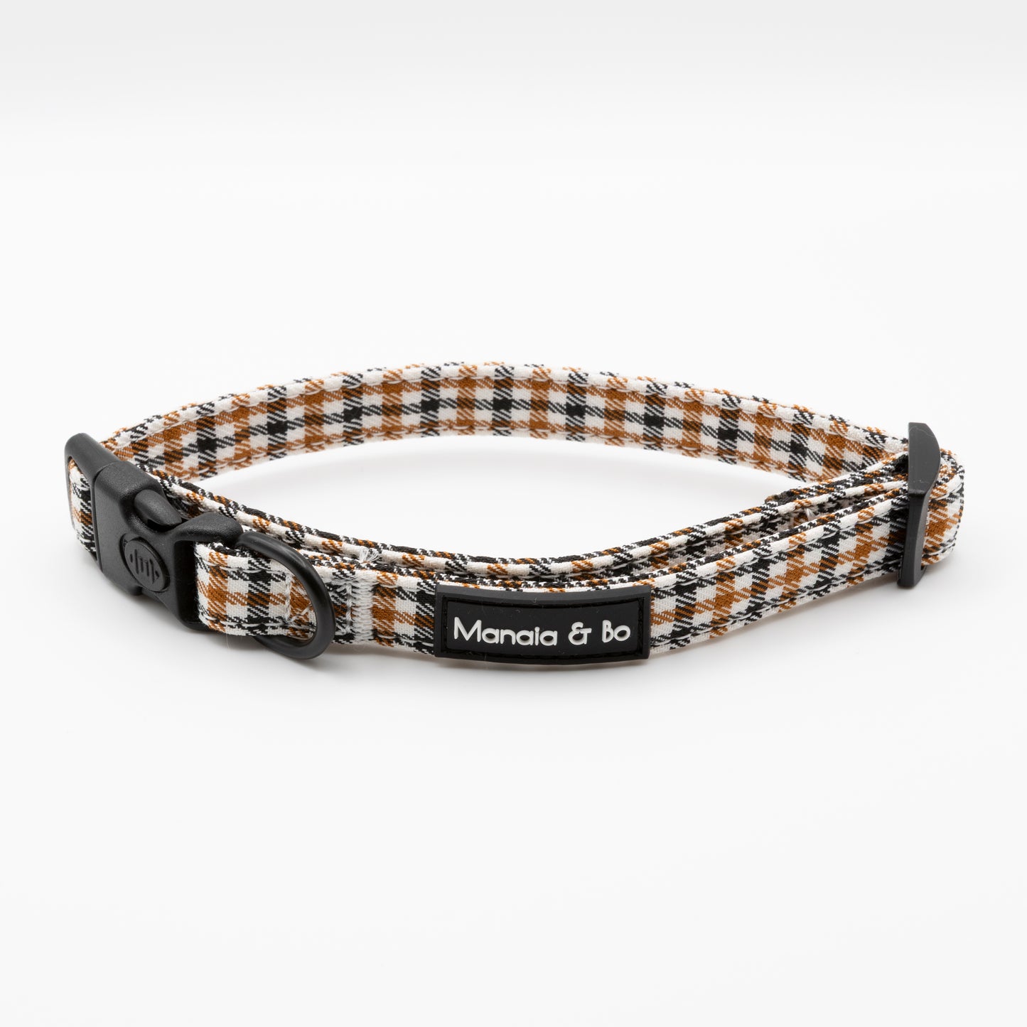 The Cocoa Heritage Lite Dog Collar features a plaid pattern in black, white, and brown, an adjustable fit with lockable buckle, metal ring, and a Manala & bo label on the side.