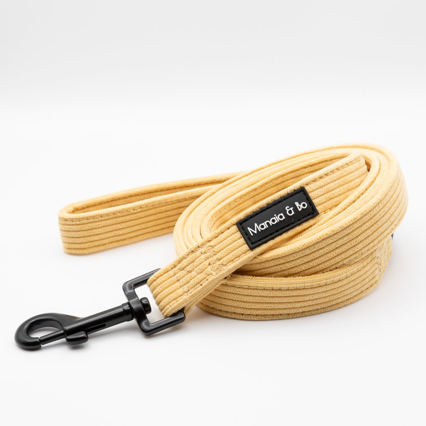 The Mellow Cord Collar & Lead is a coiled, light yellow dog leash with a metal clasp and black "Mandia & bo" label, paired with an adjustable collar, both displayed on a white background.