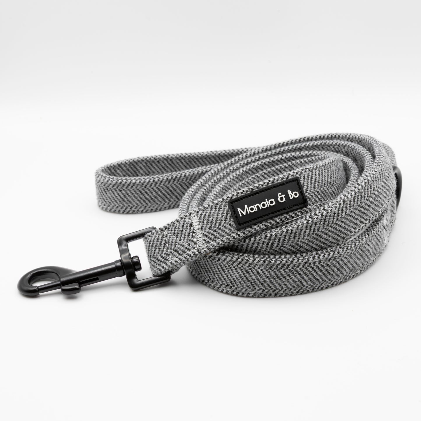 A coiled gray herringbone Monochrome Tweed Dog Lead with a black metal clasp and Manala & bo label on a white background—a stylish, durable leash designed for reliability.