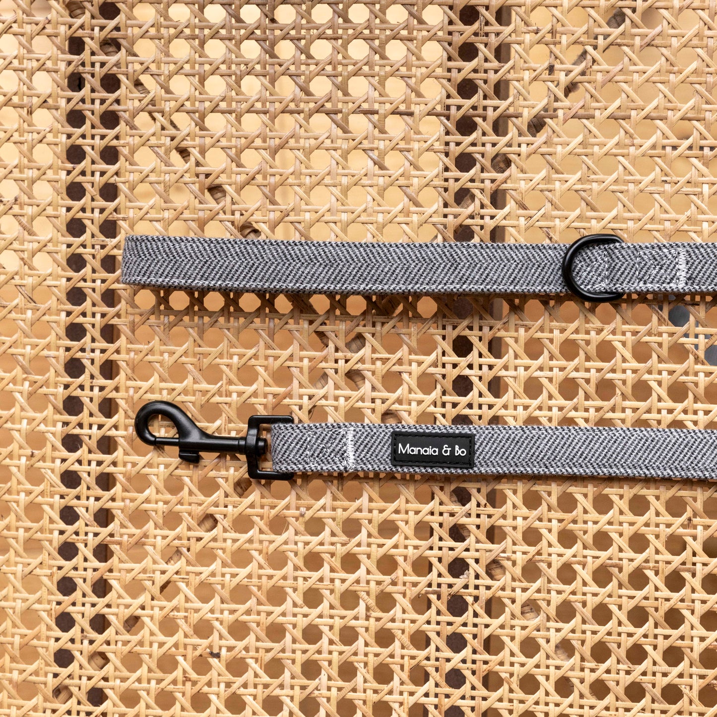 Close-up of the Monochrome Tweed Dog Lead, a gray fabric leash with black metal clips, on a woven rattan background. It’s styled with a small black label featuring white text.