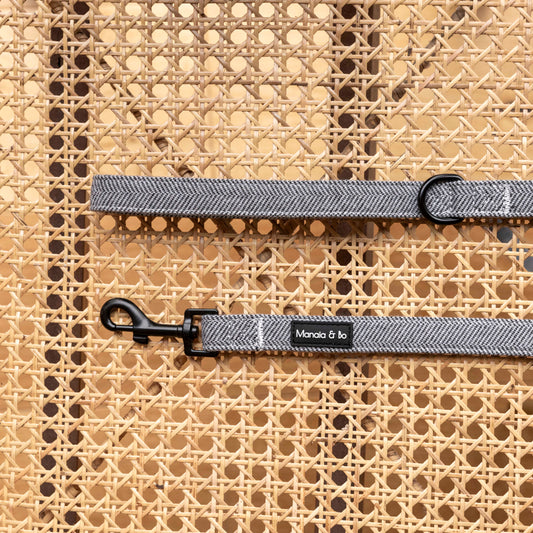 Close-up of the Monochrome Tweed Dog Lead, a gray fabric leash with black metal clips, on a woven rattan background. It’s styled with a small black label featuring white text.