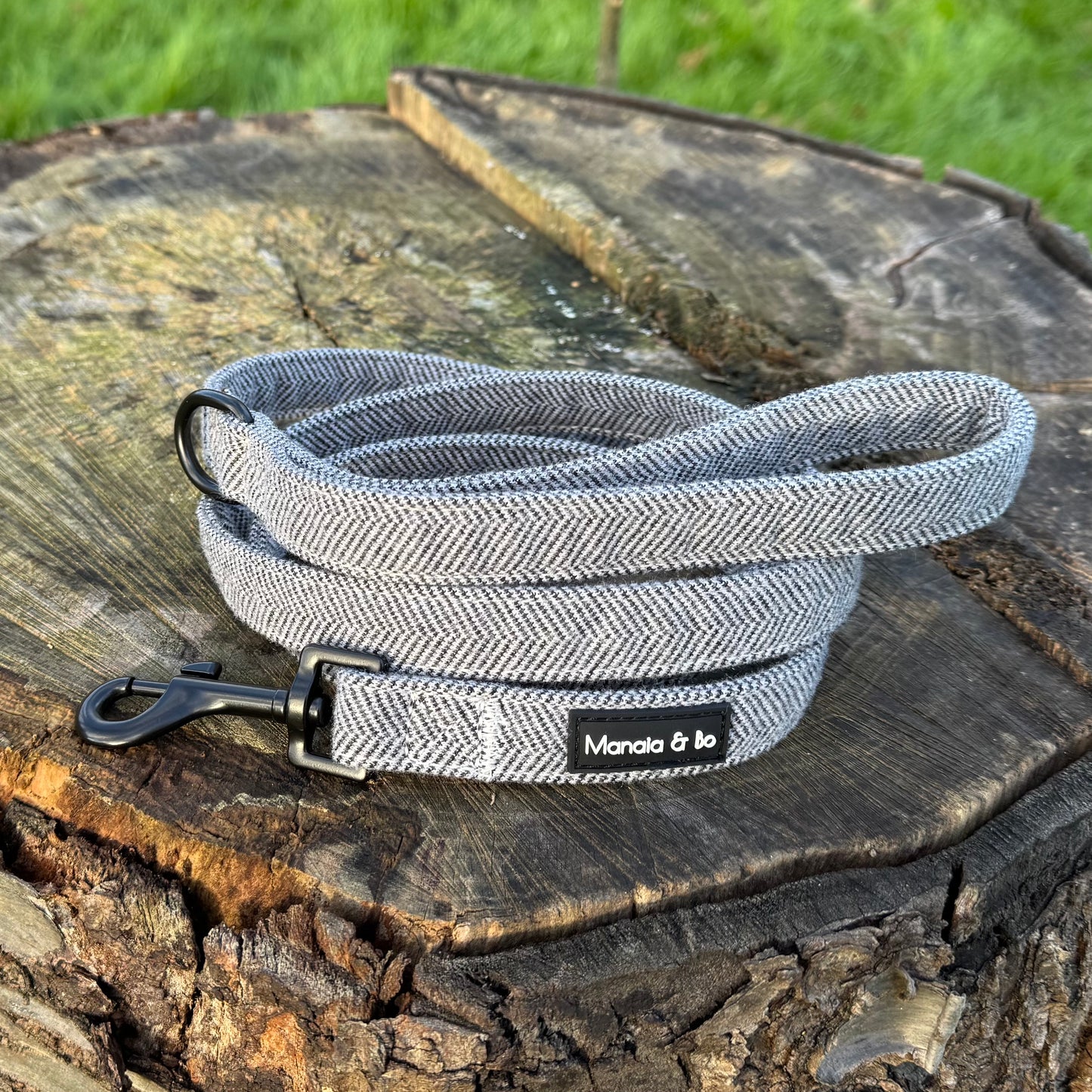 Monochrome (dark grey and white) tweed herringbone lead in a coil on top of a tree stump with grass in the background from the front. In the foreground is the black metal swivel snap hook to attach to a collar or harness