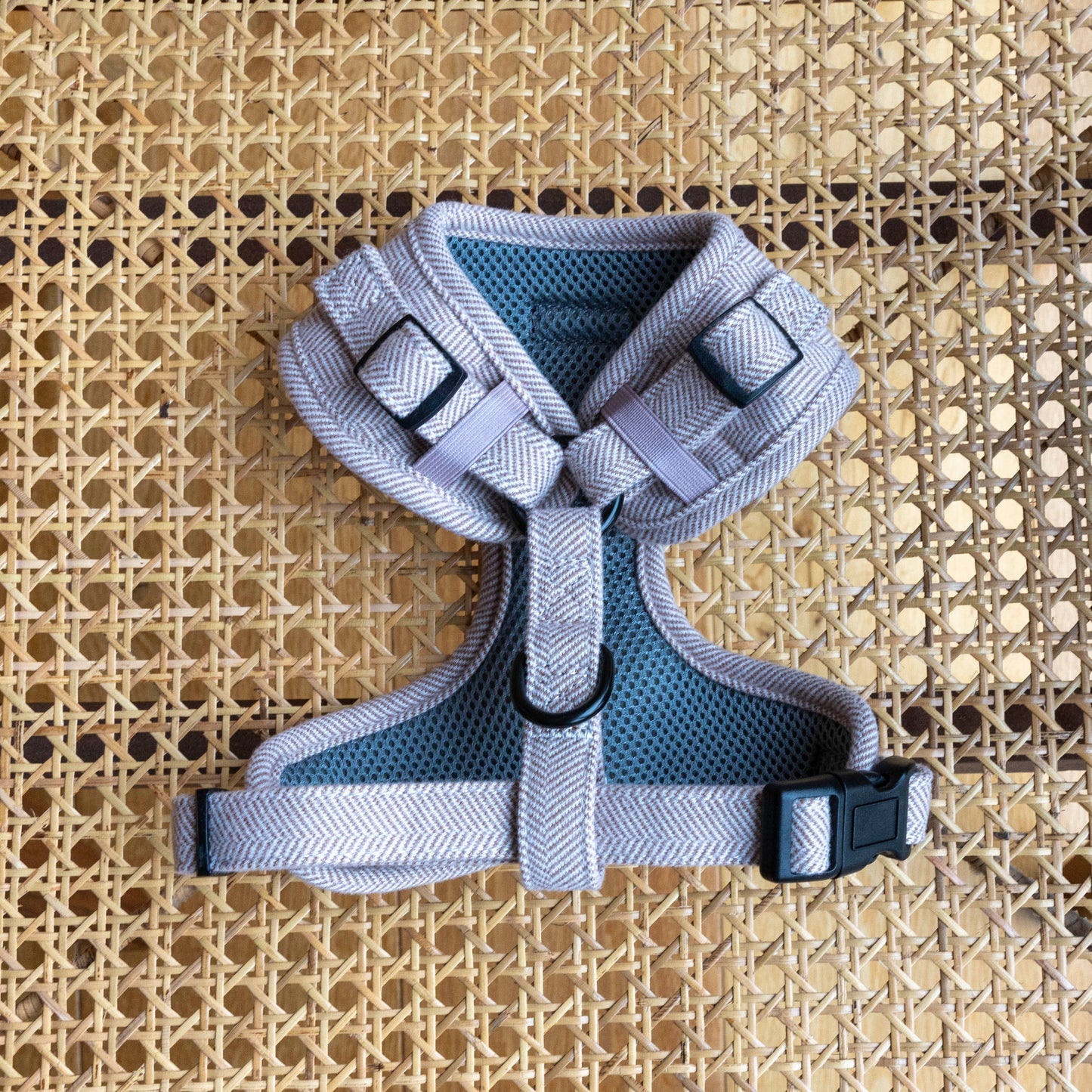 The Nougat tweed Dog Harness, featuring a beige herringbone pattern, adjustable design, black buckle and D-ring, rests on a woven rattan surface. Made from quality pet materials, it has padded straps and breathable mesh lining for your dogs comfort.