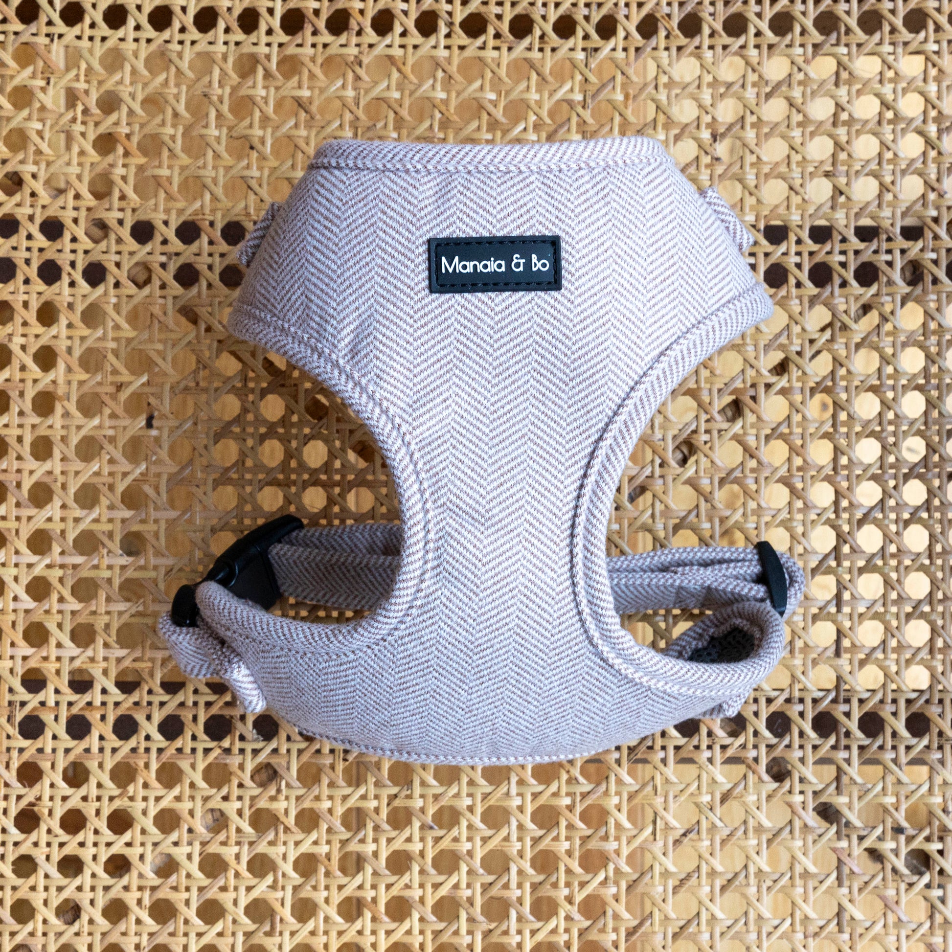 The Nougat tweed Dog Harness, made from quality materials in beige textured fabric with a black clasp, sits on a woven rattan surface. This adjustable harness offers comfort and stylish practicality for your furry friend.