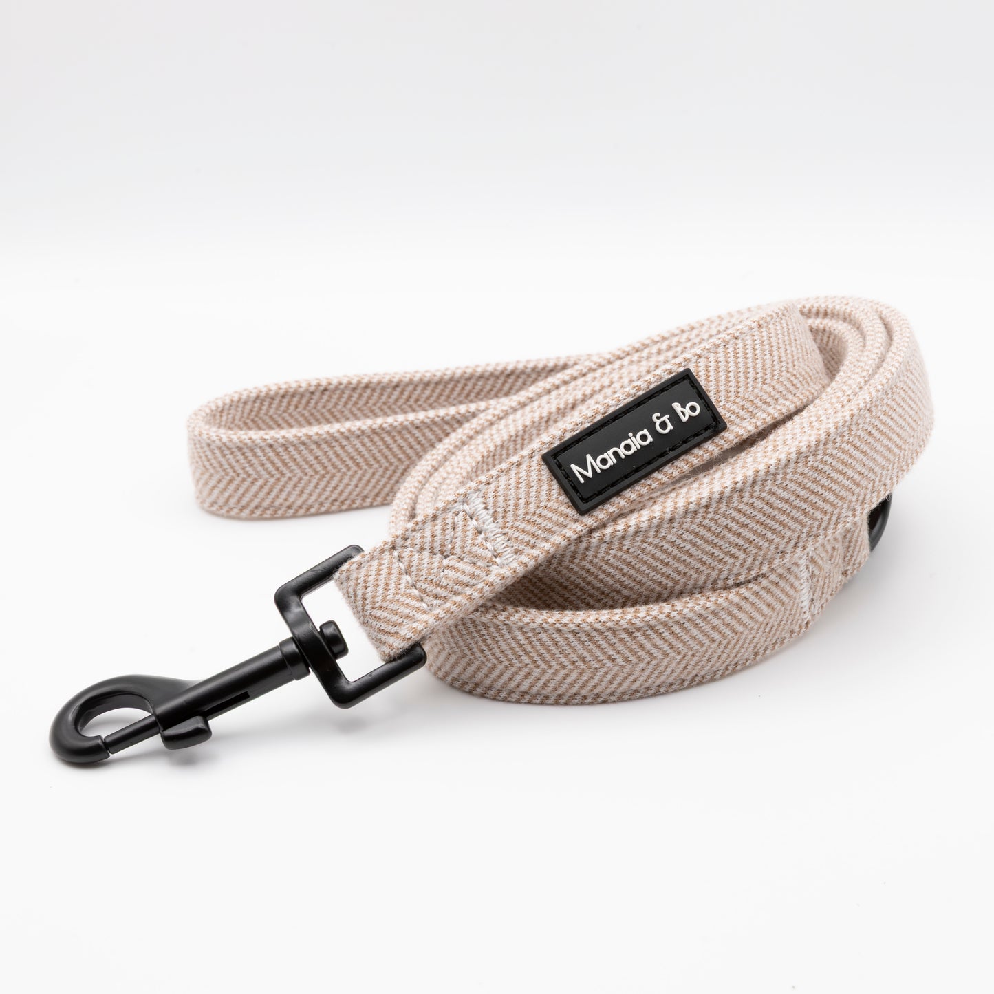 The Nougat Tweed Dog Lead, featuring a beige herringbone pattern, black metal clasp, and a black Mondo & bo label, is coiled neatly on a white background.