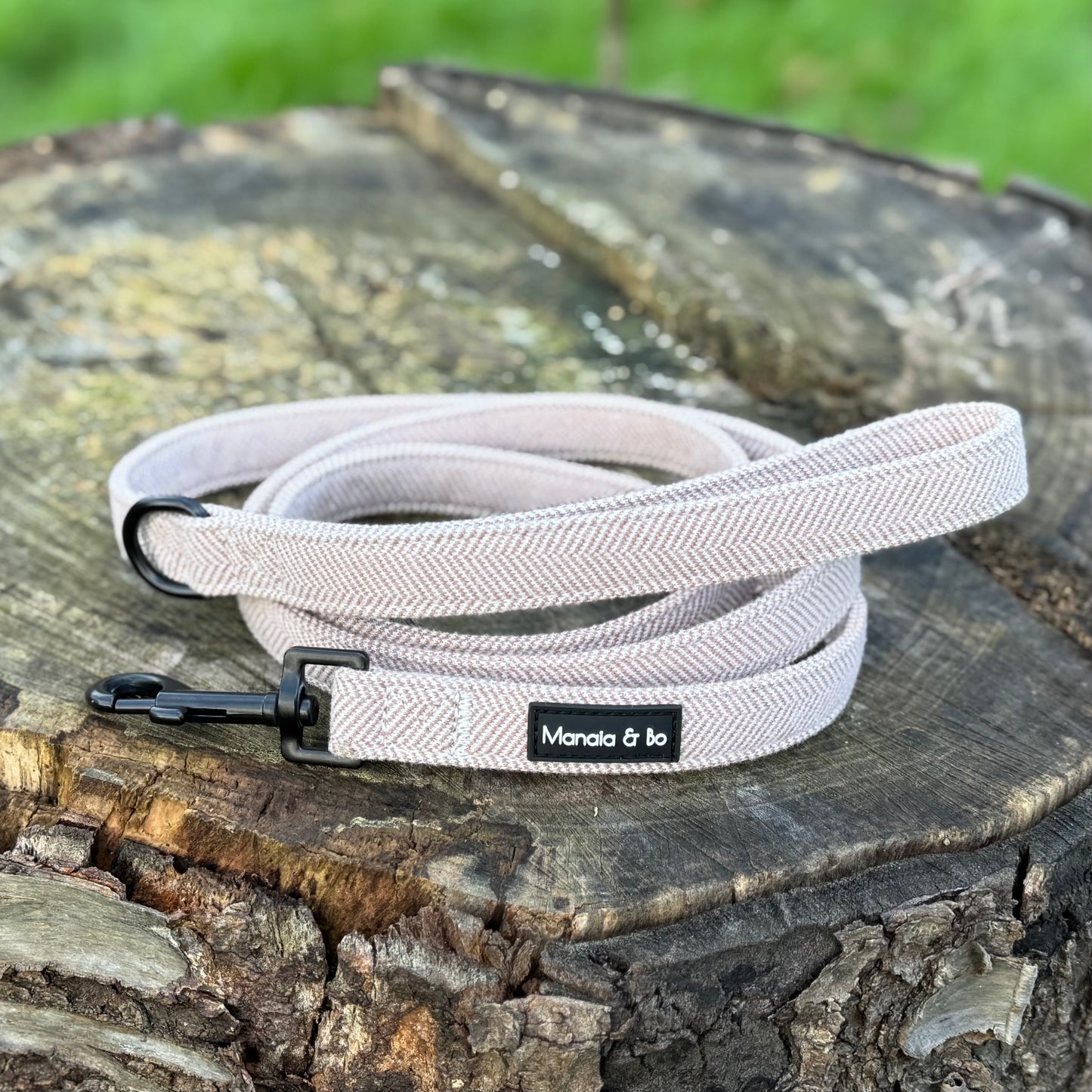 The Nougat Tweed Dog Lead, a light brown textured leash with a black clasp, exudes elegance and style as it lies neatly coiled on a tree stump.