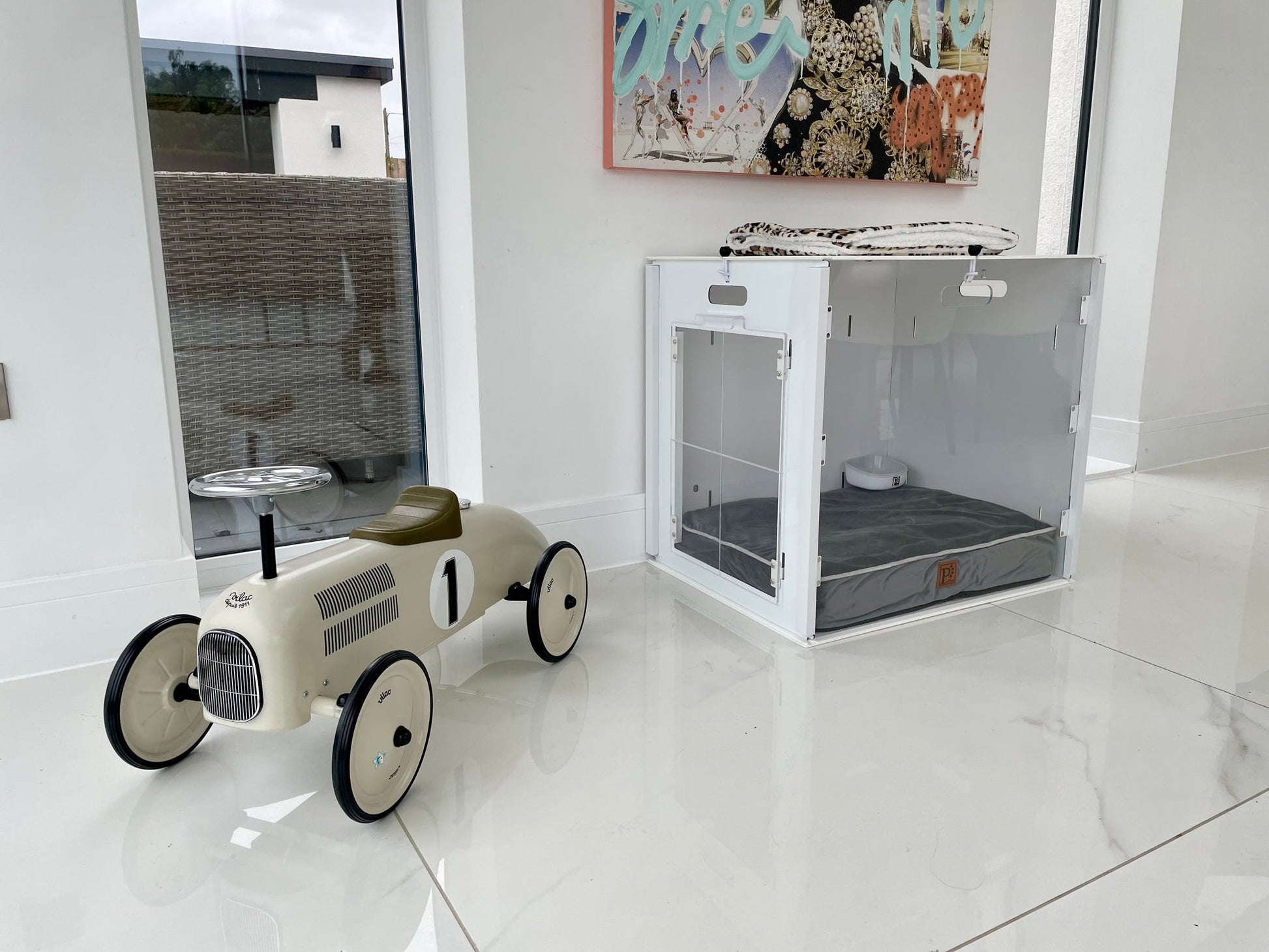 A cream-colored toy race car with the number 1 sits on a shiny white floor beside a minimalist Petpen Crate featuring a gray cushion, in a bright room with large windows.