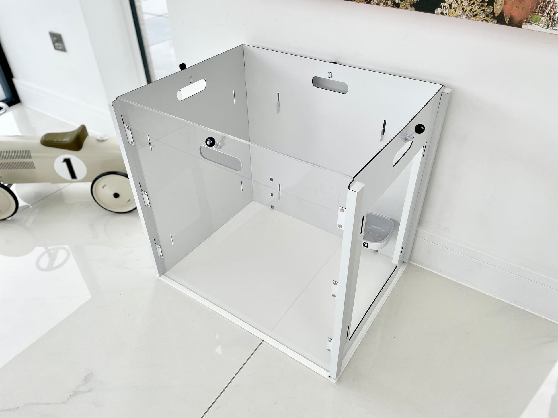 The Petpen Crate, a white and clear minimalist pet den with open sides, sits on a glossy white floor near a wall. Ideal for crate training in modern spaces, it’s stylish and practical. A small model car with the number 1 appears in the background.