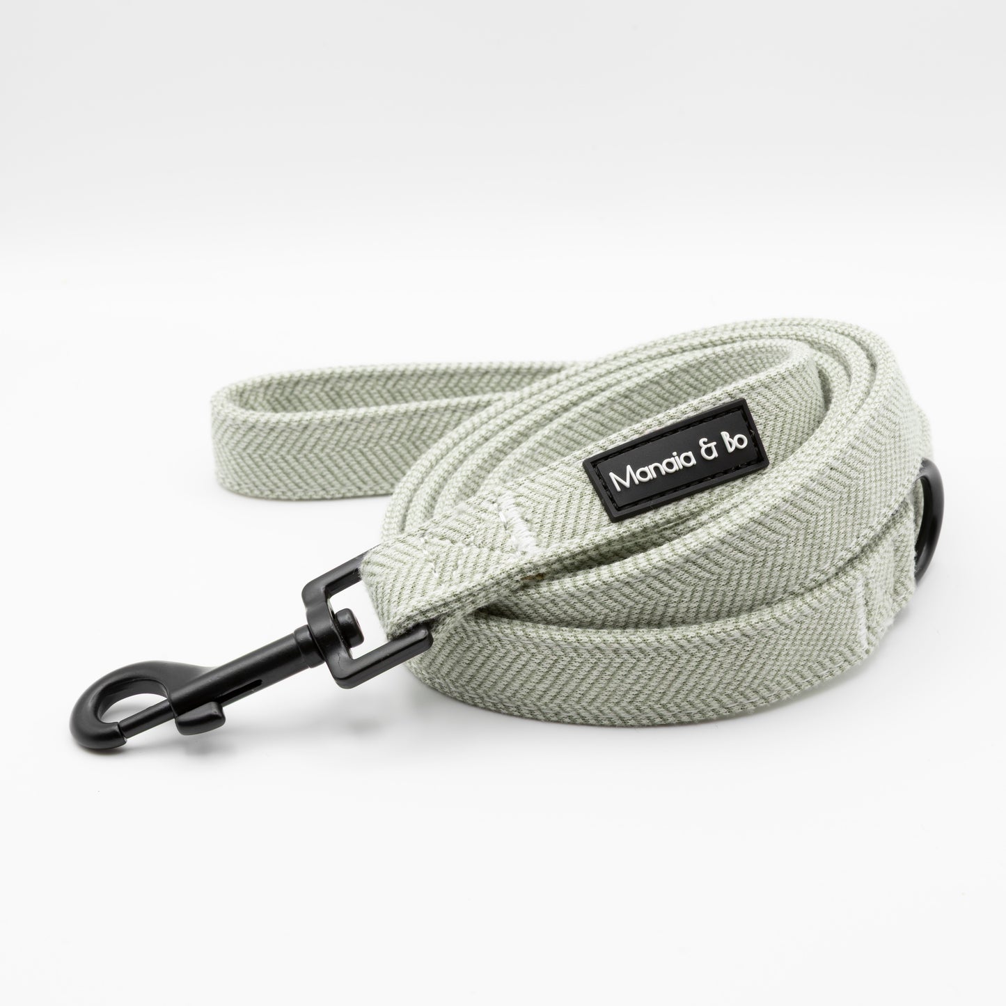 The Pine Tweed Harness & Lead features a light gray woven dog leash with a black metal clasp and a Manola & Bo label, coiled neatly on a white background—pair it with our adjustable harness for superior style and comfort.